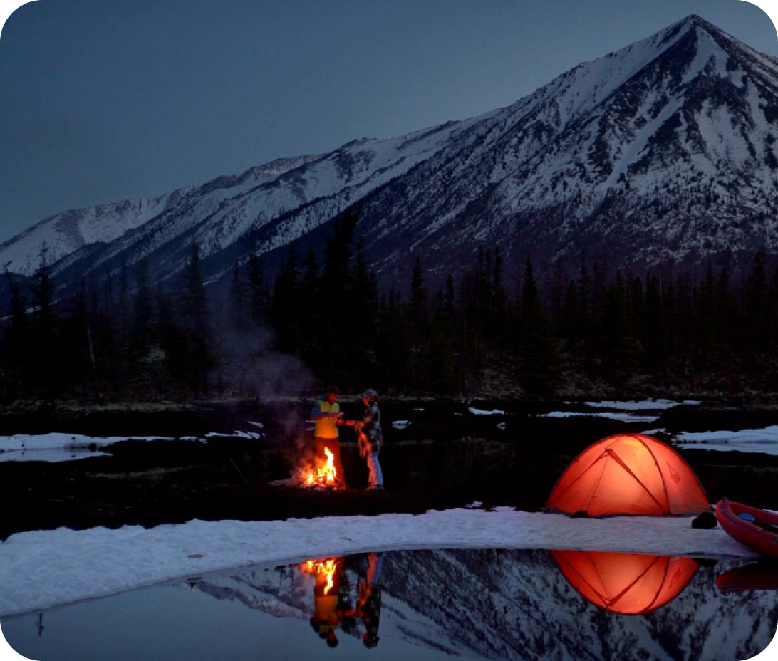 A couple camping with a fire, in a snowy mountainous setting.